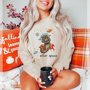 May include: A beige sweatshirt featuring a cartoon otter wearing a space helmet and flying through space. The text "otter space" is printed below the otter.