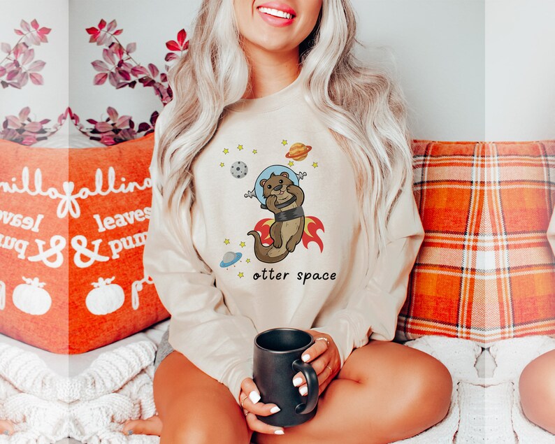 May include: A beige sweatshirt featuring a cartoon otter wearing a space helmet and flying through space. The text "otter space" is printed below the otter.