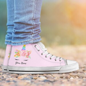 May include: Pink high-top canvas shoes with a unicorn design featuring a rainbow horn, flowers, and eyelashes. The shoes have white laces and a white rubber sole. The text "Your Name" is printed on the shoes.