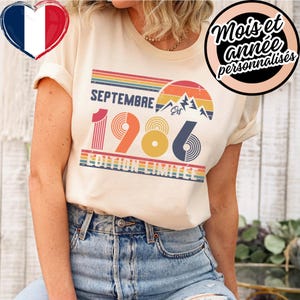 Peut inclure: Un t-shirt couleur crème arborant un graphisme de style rétro avec le texte SEPTEMBRE 1986 EDITION LIMITEE. Le motif comprend un coucher de soleil, des sommets montagneux et des rayures horizontales dans des tons orange, jaune, bleu et vert.