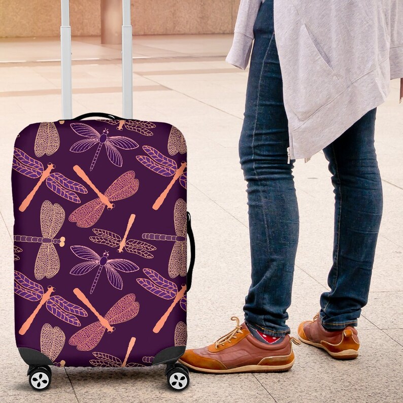 May include: Purple luggage cover with a pattern of pink and purple dragonflies. The luggage cover is on a suitcase with wheels.