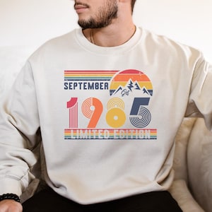 May include: A beige crewneck sweatshirt featuring a graphic print with the text September 1985 Limited Edition. The design includes a retro sunset, mountain peaks, and horizontal stripes in shades of red, orange, yellow, and blue.