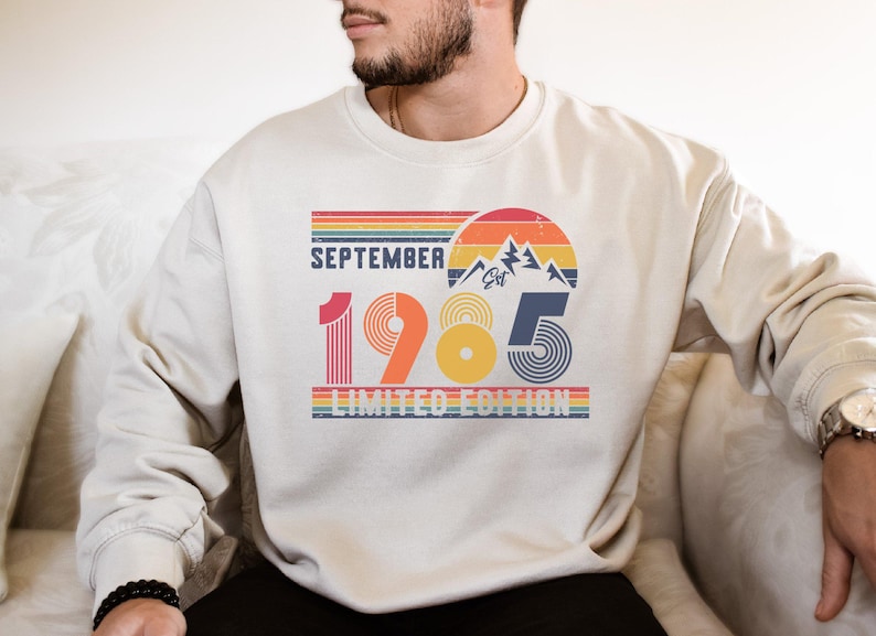 May include: A beige crewneck sweatshirt featuring a graphic print with the text September 1985 Limited Edition. The design includes a retro sunset, mountain peaks, and horizontal stripes in shades of red, orange, yellow, and blue.