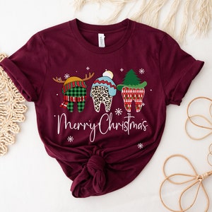 Op de afbeelding: Bordeauxrood t-shirt met een kerstthema van drie tanden. E&eacute;n tand is een rendier, een andere heeft een luipaardprint hoed en de derde is een kerstboom. De woorden "Merry Christmas" zijn in witte letters geschreven.