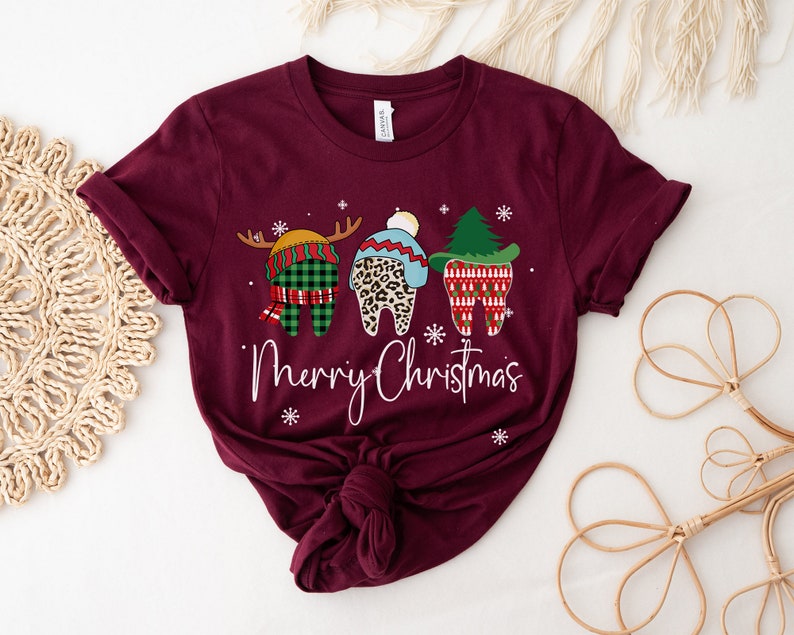 Op de afbeelding: Bordeauxrood t-shirt met een kerstthema van drie tanden. E&eacute;n tand is een rendier, een andere heeft een luipaardprint hoed en de derde is een kerstboom. De woorden "Merry Christmas" zijn in witte letters geschreven.