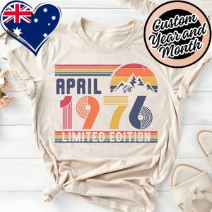 May include: Cream-colored t-shirt with a retro design featuring the text "APRIL 1976 LIMITED EDITION." The design includes a rainbow-colored stripe, a mountain graphic, and the words "Custom Year and Month" in a circular design.