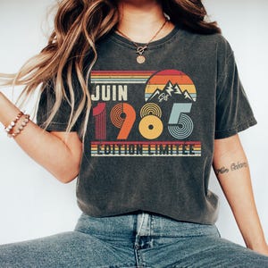 May include: A charcoal grey t-shirt featuring a retro-style graphic with the text JUIN 1985 EDITION LIMITEE. The design includes a sunset over mountains in orange, yellow, and blue stripes, with the numbers 1985 styled in bold, colourful, circular patterns.