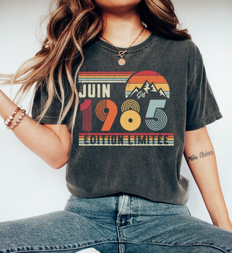 May include: A charcoal grey t-shirt featuring a retro-style graphic with the text JUIN 1985 EDITION LIMITEE. The design includes a sunset over mountains in orange, yellow, and blue stripes, with the numbers 1985 styled in bold, colourful, circular patterns.