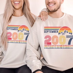 K&ouml;nnte beinhalten: Beige Sweatshirts mit einem Retro-Design mit dem Text "SEPTEMBER 2007 LIMITED EDITION" in einer farbenfrohen, geschichteten Schriftart. Das Design beinhaltet eine Berg-Grafik und einen Regenbogenbogen.