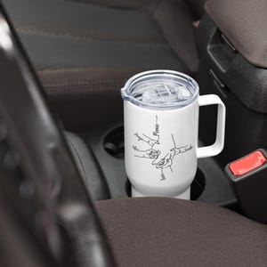 May include: White travel mug with a handle and a clear lid, featuring a black line drawing of hands holding each other. The names "Hugo", "Alex", "Lola", "Mamie", and "Sasha" are written on the mug.