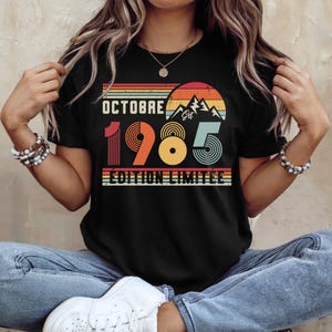 May include: A black short-sleeve t-shirt featuring a retro-style graphic with the text "OCTOBRE 1985 &Eacute;DITION LIMIT&Eacute;E". The design includes a sunset, mountain silhouette, and horizontal stripes in shades of orange, yellow, and teal.