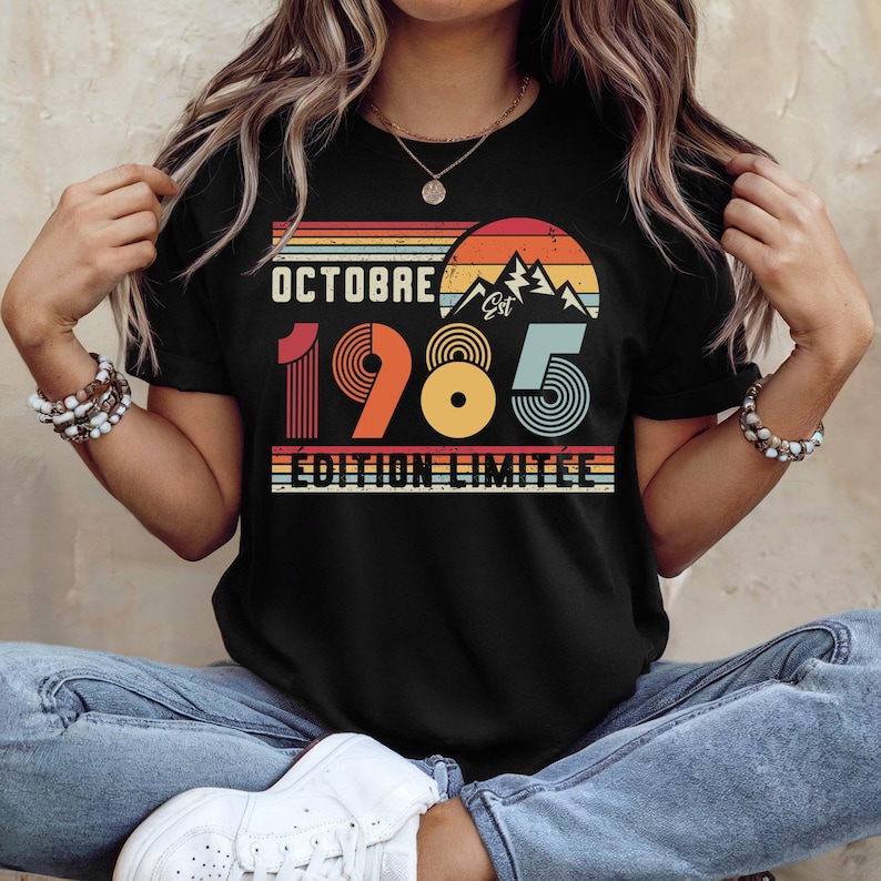 May include: A black short-sleeve t-shirt featuring a retro-style graphic with the text "OCTOBRE 1985 &Eacute;DITION LIMIT&Eacute;E". The design includes a sunset, mountain silhouette, and horizontal stripes in shades of orange, yellow, and teal.