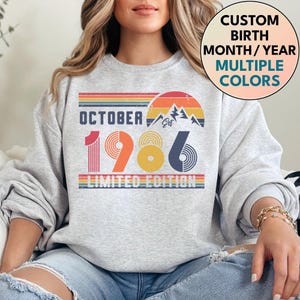 May include: A light gray crewneck sweatshirt featuring a retro-style graphic with the text OCTOBER 1986 LIMITED EDITION. The design includes a sunset, mountain peaks, and horizontal stripes in orange, yellow, and blue tones. A circular badge reads CUSTOM BIRTH MONTH/YEAR MULTIPLE COLORS.
