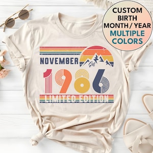 May include: A cream-colored t-shirt featuring a retro-style graphic with the text NOVEMBER 1986 LIMITED EDITION. The design includes a sunset, mountain silhouette, and horizontal stripes in red, orange, yellow, and blue. A circular badge reads CUSTOM BIRTH MONTH/YEAR MULTIPLE COLORS.