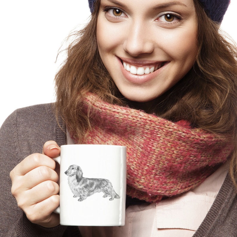 Puede incluir: Una taza de cer&aacute;mica blanca con una ilustraci&oacute;n en blanco y negro de un perro salchicha. La taza est&aacute; siendo sostenida por una persona que lleva una bufanda rosa y un gorro azul.