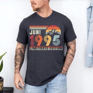 May include: Dark gray t-shirt with a retro design featuring the word "JUNI" above "1995" and "LIMITIERTE EDITION". The design includes a mountain scene and a sunset in shades of orange, red, and yellow. The shirt is a crew neck.
