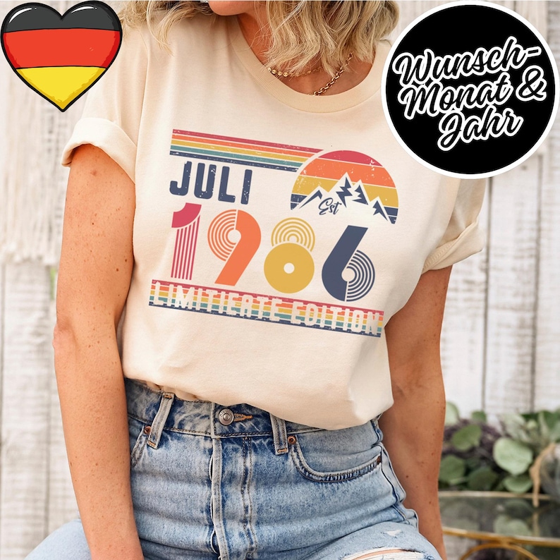 May include: A cream-colored t-shirt featuring a retro-style graphic with the text JULI 1986, a mountain illustration, and the words LIMITIERTE EDITION. The design includes horizontal stripes in orange, yellow, and blue, with a sunset motif.