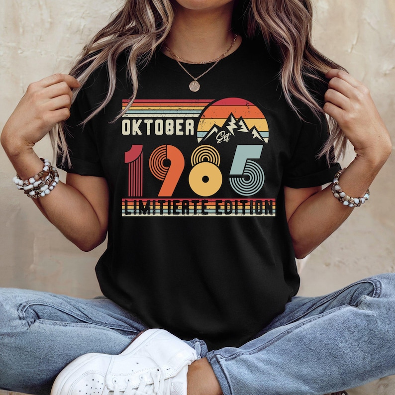 May include: A black t-shirt featuring a retro-style graphic with a sunset, mountain silhouette, and the text "OKTOBER 1985 LIMITIERTE EDITION". The design uses horizontal stripes in orange, yellow, and teal, with the numbers 1985 in large font.