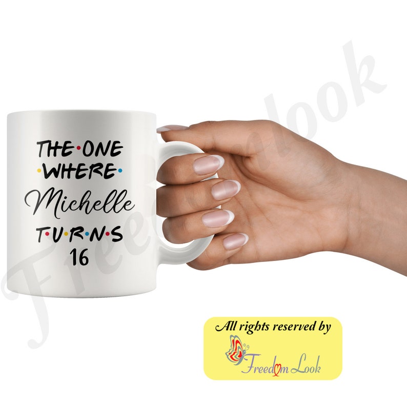 K&ouml;nnte beinhalten: Eine wei&szlig;e Keramiktasse mit schwarzem Text, der "The One Where Michelle Turns 16" lautet. Der Text ist in einer Schriftart &auml;hnlich dem Friends-Logo. Die Tasse hat einen schwarzen Henkel und wird von einer Hand gehalten.