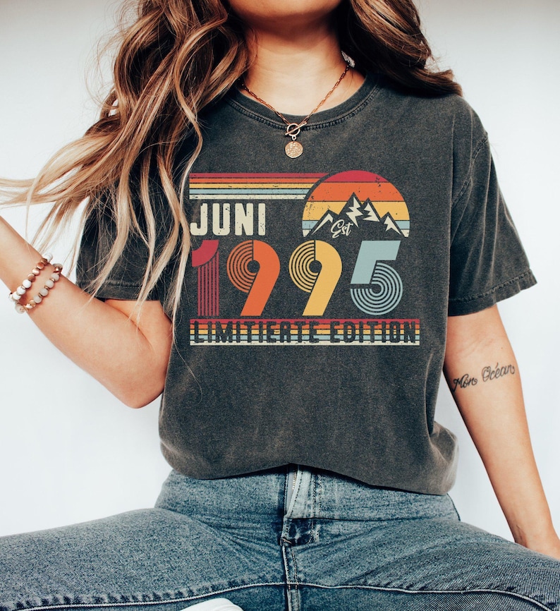 May include: Dark gray t-shirt with a retro design featuring the text "JUNI 1995 LIMITIERTE EDITION" in a colorful, vintage style. The design includes a mountain graphic and a sunset-like background, evoking a nostalgic feel.