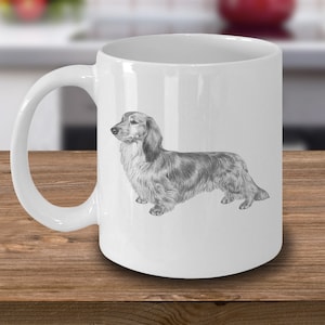 Puede incluir: Una taza de caf&eacute; de cer&aacute;mica blanca con un dibujo a l&aacute;piz en blanco y negro de un dachshund de pelo largo.