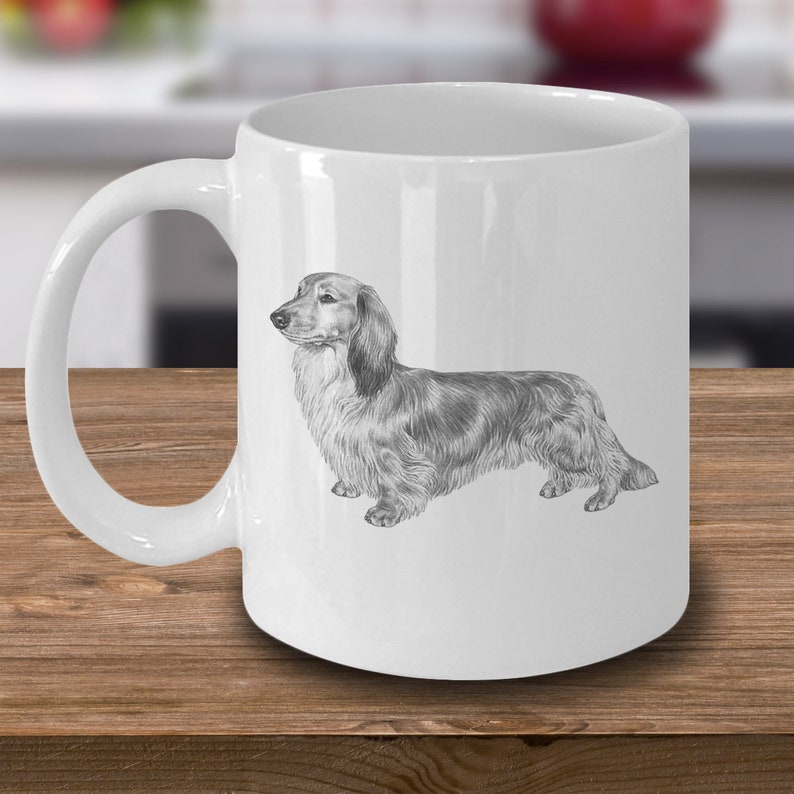 Puede incluir: Una taza de caf&eacute; de cer&aacute;mica blanca con un dibujo a l&aacute;piz en blanco y negro de un dachshund de pelo largo.