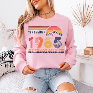 May include: A light pink crewneck sweatshirt featuring a retro-style graphic with the text SEPTEMBER 1985 LIMITED EDITION. The design includes a sunset, mountain peaks, and horizontal stripes in shades of orange, yellow, blue, and green.
