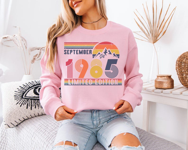 May include: A light pink crewneck sweatshirt featuring a retro-style graphic with the text SEPTEMBER 1985 LIMITED EDITION. The design includes a sunset, mountain peaks, and horizontal stripes in shades of orange, yellow, blue, and green.