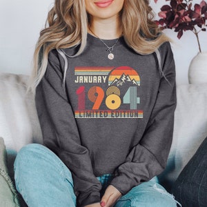 May include: Dark gray sweatshirt with a retro design featuring the text "JANUARY 1984 LIMITED EDITION." The graphic includes a mountain scene and a sunset design in orange, yellow, and teal. The sweatshirt is a crew neck style.