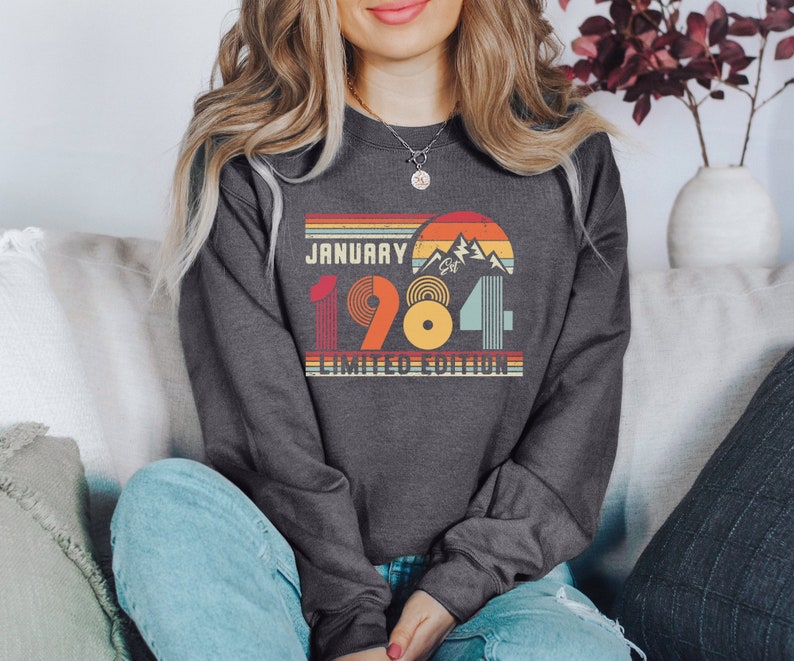 May include: Dark gray sweatshirt with a retro design featuring the text "JANUARY 1984 LIMITED EDITION." The graphic includes a mountain scene and a sunset design in orange, yellow, and teal. The sweatshirt is a crew neck style.