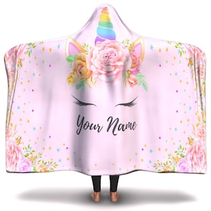 Pode incluir: Cobertor rosa com capuz com um design de unicórnio. O unicórnio tem um chifre arco-íris, uma coroa de flores e os olhos fechados. O cobertor tem um padrão de glitter dourado e o texto "Your Name" está impresso na frente.