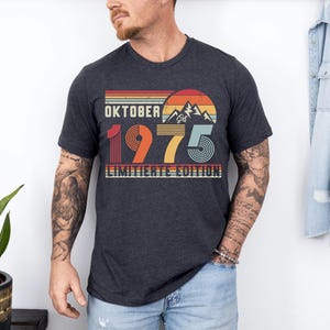 May include: A dark heather gray short-sleeve t-shirt featuring a retro-style graphic with a mountain silhouette, a sun, and the text "OKTOBER 1975 LIMITIERTE EDITION" in a multi-colored, striped design with red, orange, yellow, and blue tones.