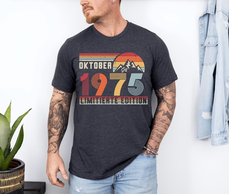 May include: A dark heather gray short-sleeve t-shirt featuring a retro-style graphic with a mountain silhouette, a sun, and the text "OKTOBER 1975 LIMITIERTE EDITION" in a multi-colored, striped design with red, orange, yellow, and blue tones.