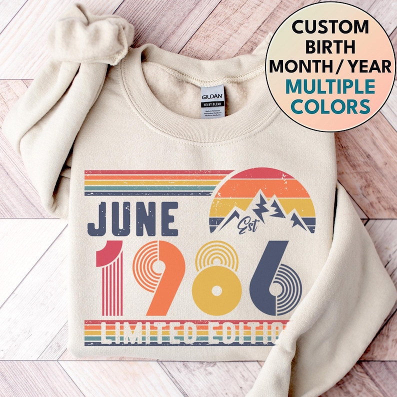 May include: Cream-colored sweatshirt with a retro design featuring the text "JUNE 1986" in a vintage font. The design includes a rainbow, mountains, and the words "LIMITED EDITION". The sweatshirt also has the text "CUSTOM BIRTH MONTH/YEAR MULTIPLE COLORS".