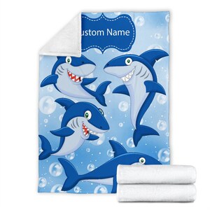 May include: A blue fleece blanket with a cartoon design of four sharks swimming in a blue ocean with white bubbles. The blanket has a blue banner with the text "Custom Name" in white.