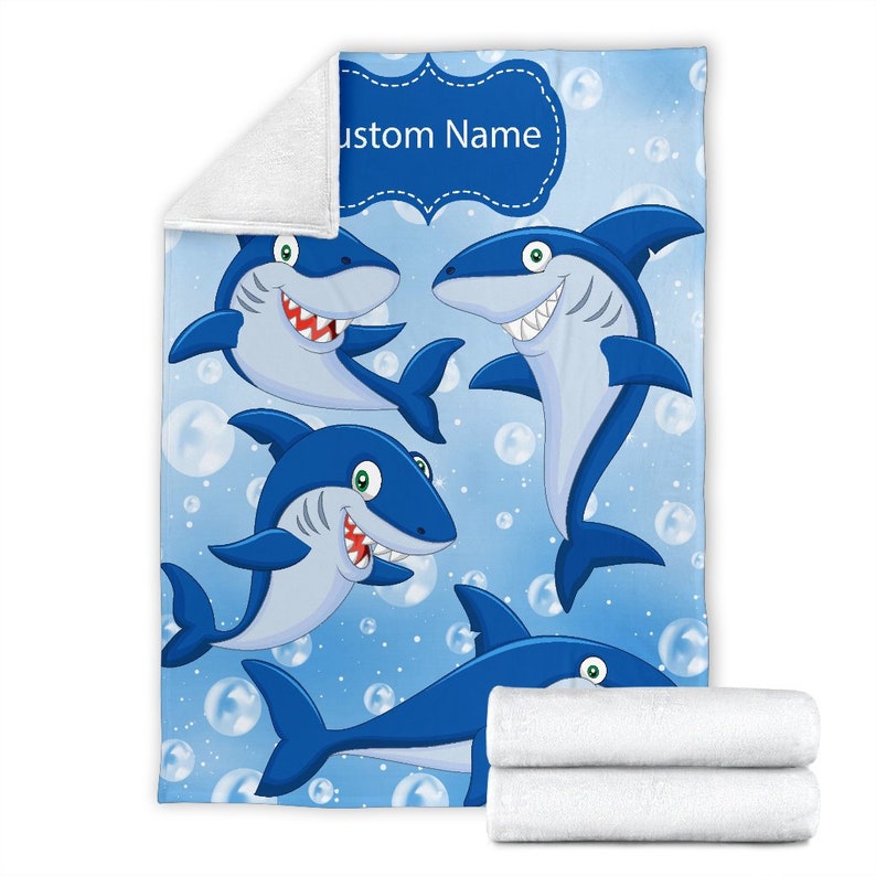 May include: A blue fleece blanket with a cartoon design of four sharks swimming in a blue ocean with white bubbles. The blanket has a blue banner with the text "Custom Name" in white.
