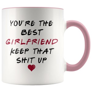 May include: White ceramic coffee mug with a pink rim and handle. The mug has black text that reads "You're the best girlfriend keep that sh*t up" with a red heart at the bottom.