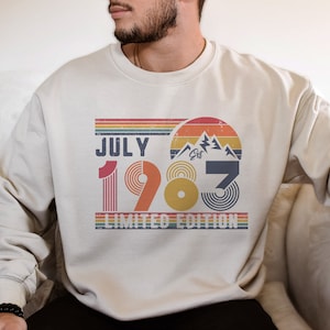 May include: Cream-colored sweatshirt with a retro design featuring the text "JULY 1983 LIMITED EDITION." The design includes a mountain graphic, rainbow stripes, and a vintage aesthetic. A stylish and comfortable apparel item.