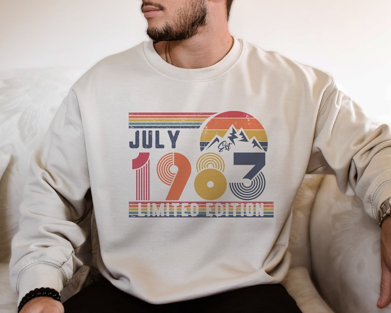 May include: Cream-colored sweatshirt with a retro design featuring the text "JULY 1983 LIMITED EDITION." The design includes a mountain graphic, rainbow stripes, and a vintage aesthetic. A stylish and comfortable apparel item.