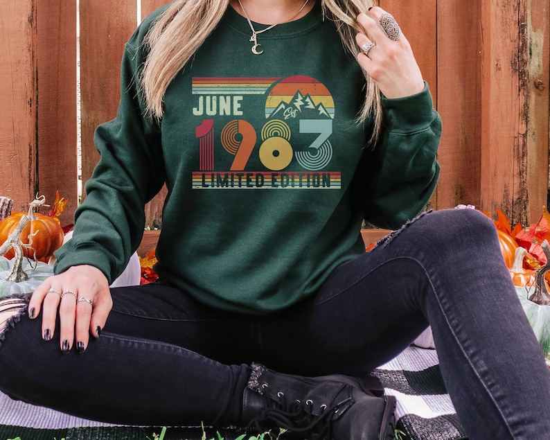 May include: Dark green sweatshirt with the text "JUNE 1983 LIMITED EDITION" in a retro design. The design includes a mountain range and a sunset color scheme. The sweatshirt is a long-sleeved crew neck.