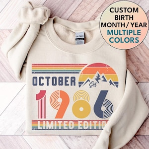 Op de afbeelding: Een crèmekleurig sweatshirt met ronde hals en een grafische print met de tekst OCTOBER 1986 LIMITED EDITION. Het ontwerp bevat een retro zonsondergang, bergsilhouetten en horizontale strepen in de kleuren rood, oranje, geel en blauw.