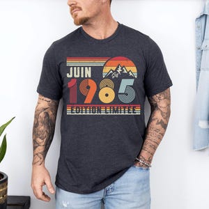May include: A dark heather grey short-sleeve t-shirt featuring a retro-style graphic with a sunset, mountain silhouette, and the text "JUIN 1985 EDITION LIMITEE" in a multi-coloured design with horizontal stripes in red, orange, and blue tones.