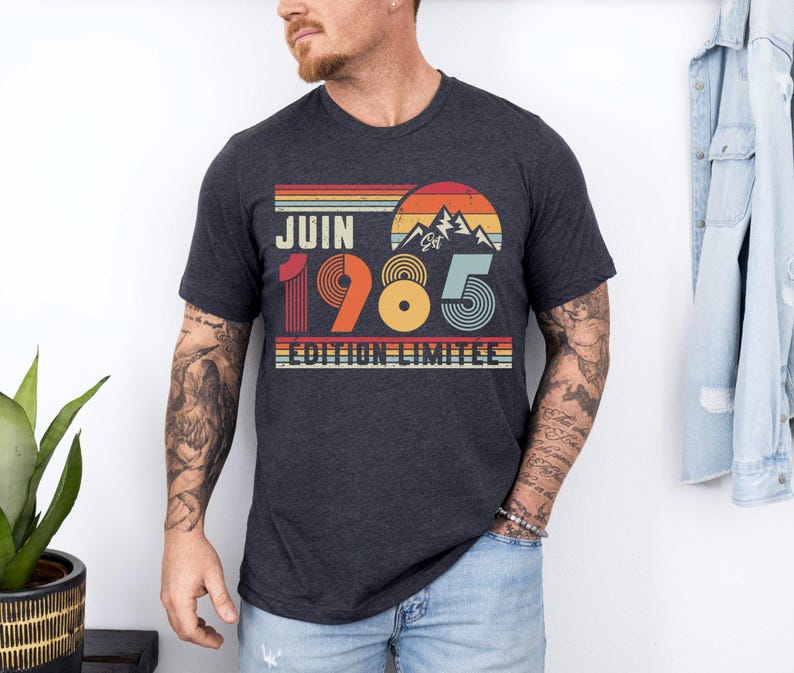 May include: A dark heather grey short-sleeve t-shirt featuring a retro-style graphic with a sunset, mountain silhouette, and the text "JUIN 1985 EDITION LIMITEE" in a multi-coloured design with horizontal stripes in red, orange, and blue tones.