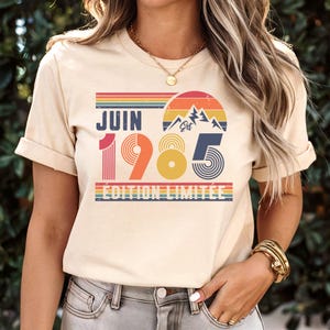 May include: A cream-coloured short-sleeve t-shirt featuring a graphic print with the text JUIN 1985 &Eacute;DITION LIMIT&Eacute;E. The design includes a retro-style sunset, mountain silhouette, and horizontal stripes in shades of red, orange, yellow, and blue.