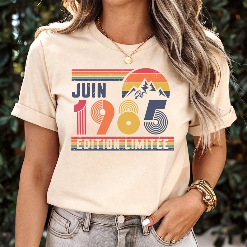 May include: A cream-coloured short-sleeve t-shirt featuring a graphic print with the text JUIN 1985 &Eacute;DITION LIMIT&Eacute;E. The design includes a retro-style sunset, mountain silhouette, and horizontal stripes in shades of red, orange, yellow, and blue.