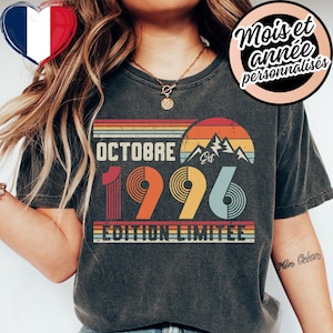 Może przedstawiać: Ciemnoszary t-shirt z grafiką w stylu retro, przedstawiającą poziome paski, sylwetkę góry oraz napis OCTOBRE 1996 EDITION LIMITEE. Wzór zawiera motyw zachodzącego słońca w odcieniach pomarańczu, żółci i jasnego błękitu.