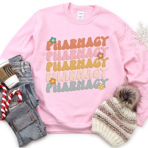 May include: Pink sweatshirt with the word "Pharmacy" repeated five times in a rainbow of colors with a flower design. The sweatshirt is laid out on a white surface with a pair of jeans, a coffee cup, and a winter hat.