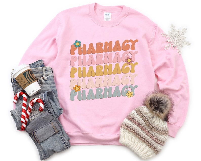 May include: Pink sweatshirt with the word "Pharmacy" repeated five times in a rainbow of colors with a flower design. The sweatshirt is laid out on a white surface with a pair of jeans, a coffee cup, and a winter hat.