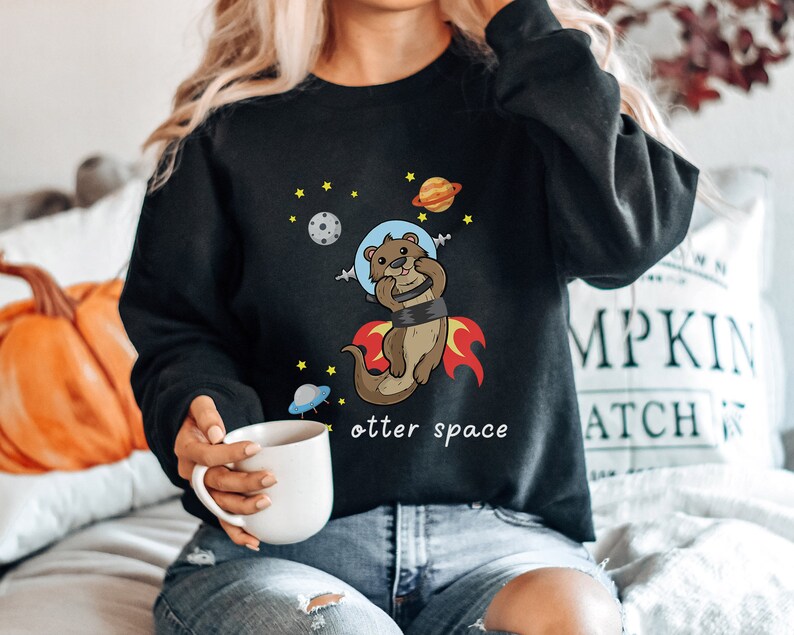 May include: Black sweatshirt featuring a cartoon otter wearing a space helmet and flying through space. The text "otter space" is printed below the otter.