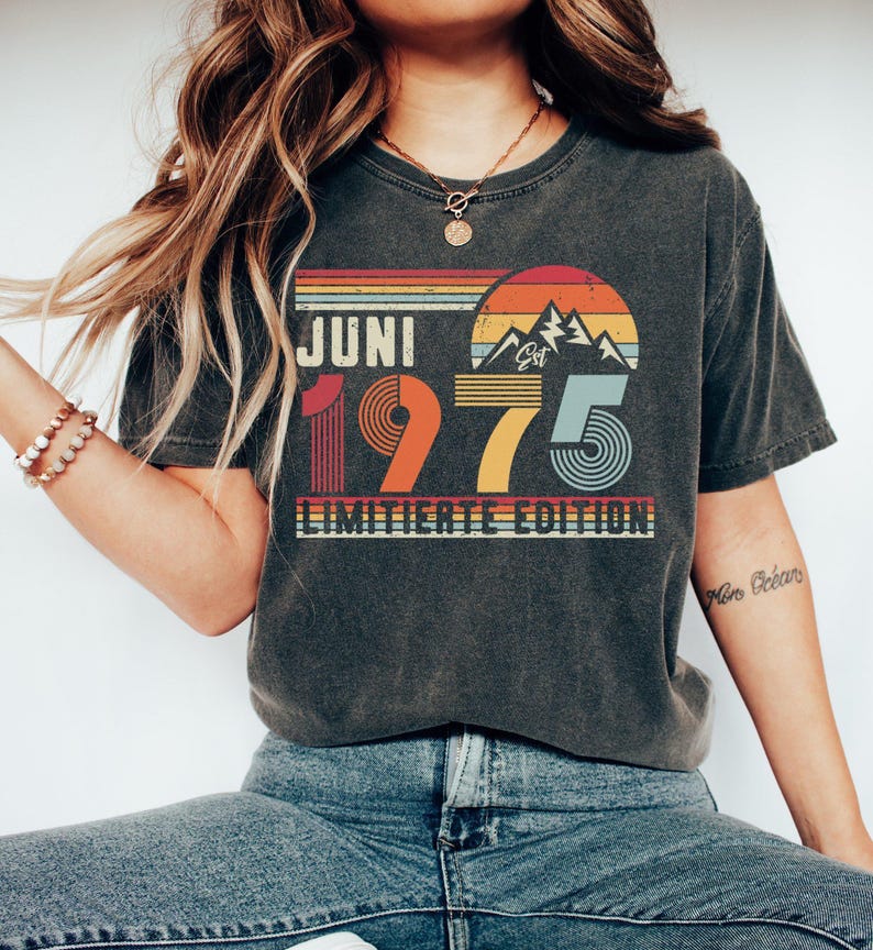 May include: A charcoal gray short-sleeve t-shirt featuring a retro-style graphic with the text JUNI 1975 LIMITIERTE EDITION. The design includes a sunset over mountain peaks in shades of orange, yellow, and blue, with striped horizontal lines.